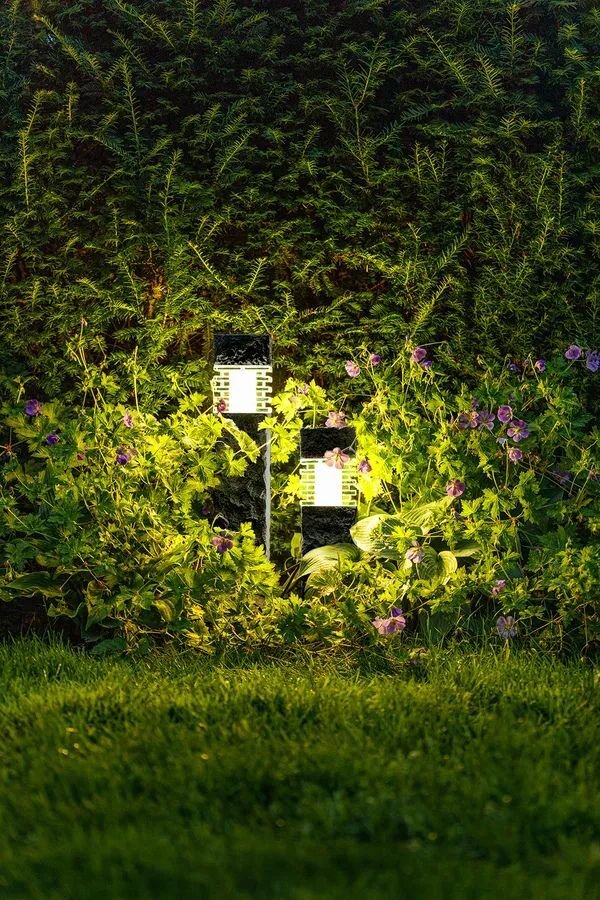 Deux bornes lumineuses de jardin aspect pierre allumées, éclairage d’un massif végétal devant une h